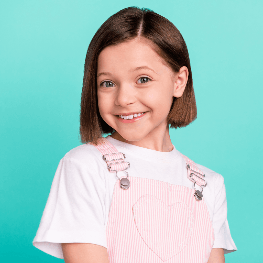 A cheerful young girl with a neat, chin-length bob haircut smiles brightly against a turquoise background. Dressed in a white t-shirt and pink striped dungarees, she looks fresh and happy—capturing the fun, friendly atmosphere of children’s haircut services at Honeys Hair & Beauty in Taunton.