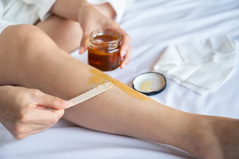 Person applying warm wax to lower leg at home using a wooden spatula, with waxing supplies in the background — representing DIY waxing.