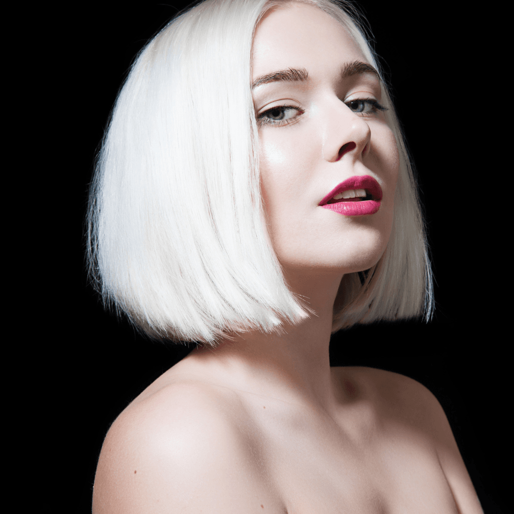 A striking portrait of a woman with a sleek, platinum blonde bob against a black background. Her hair is smooth and glossy, styled to perfection, and paired with flawless makeup and bold pink lipstick. The image showcases professional hair colouring and modern styling, reflecting the premium services offered at Honeys Hair & Beauty in Taunton.