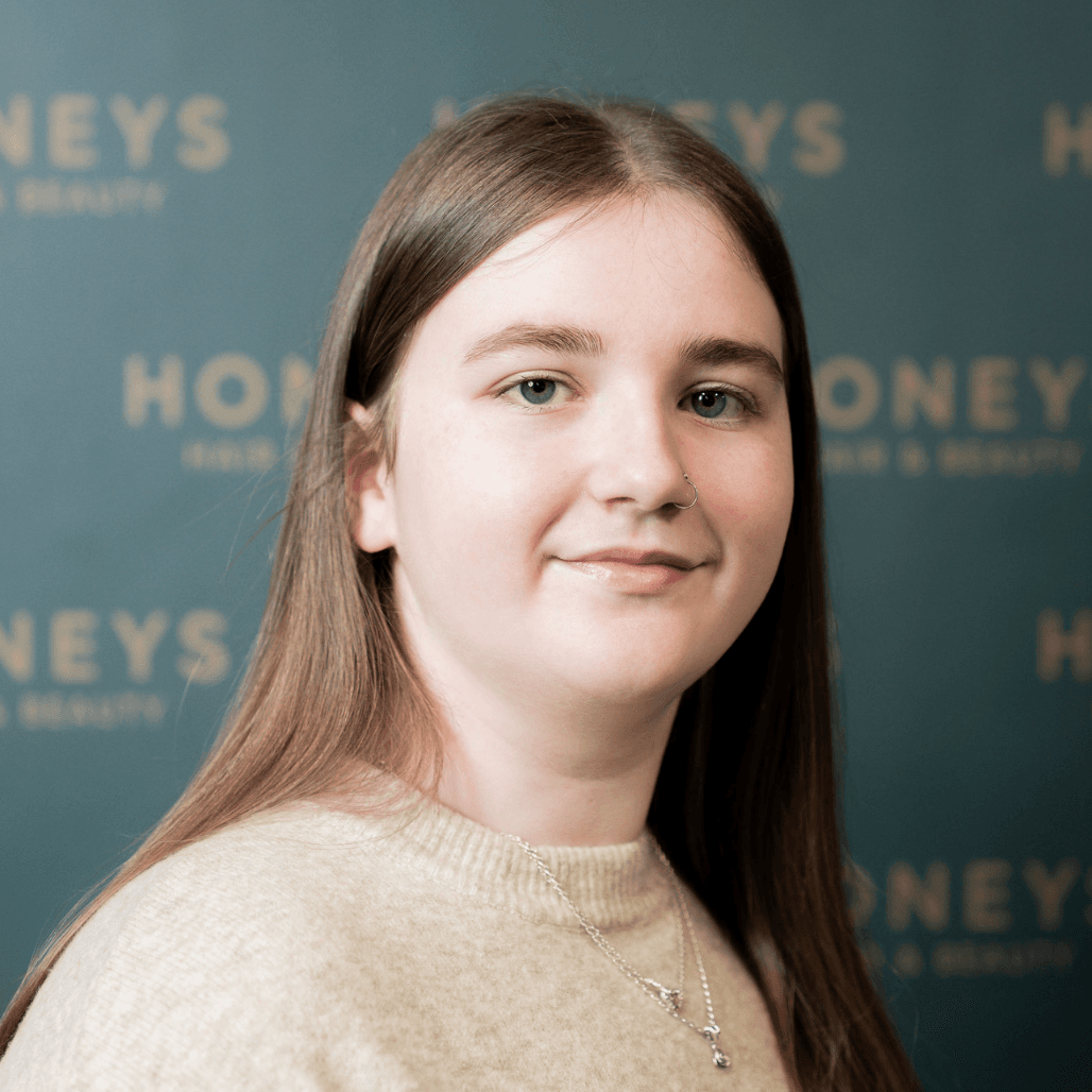 Mia, with straight light brown hair and a gentle smile, stands confidently in front of the Honeys Hair & Beauty branding. Her soft, approachable look reflects the relaxed and friendly team environment at the Taunton salon.